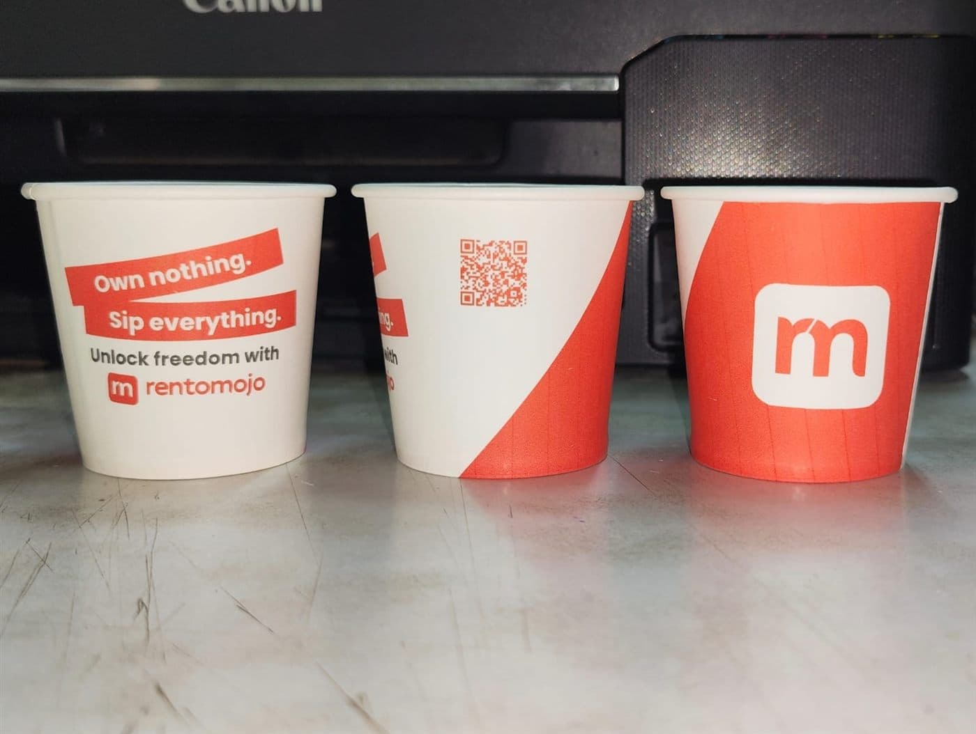 RentoMojo branded cups on a printer stand showing three cup designs including the QR code and logo variant
